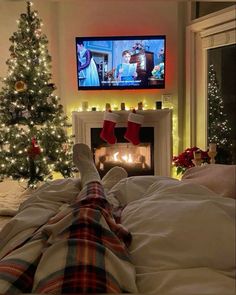 a person laying in bed with their feet on the pillow watching tv next to a christmas tree