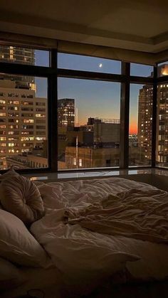 an unmade bed in front of a large window overlooking the city at night time