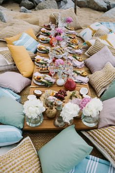 a table set up with food and pillows on the beach for an outdoor dinner party