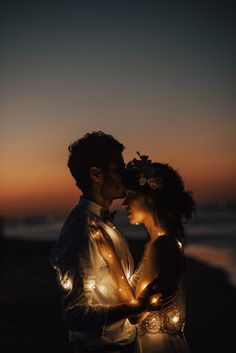 a man and woman standing next to each other near the ocean at night with lights in their hair