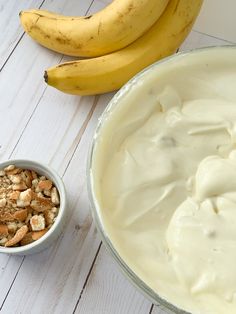 a bowl of yogurt next to two bananas on a white wooden table,