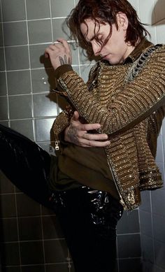 a man in black leather pants and a gold jacket sitting on a tiled bathroom wall