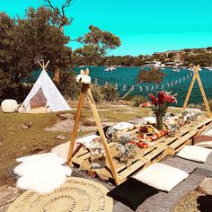 a picnic table set up with food and pillows on the grass near water in front of a teepee tent