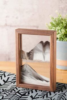 a small wooden frame with an image of mountains in the middle on a table next to a potted plant