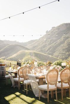 an outdoor dining table set up with white linens and wooden chairs in front of mountains