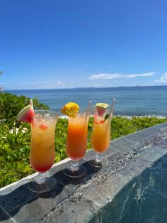 three drinks sitting on top of a table next to the ocean in front of some bushes