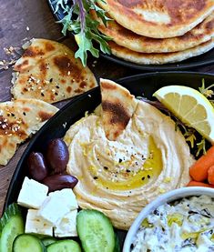 two black plates filled with hummus, cucumber, carrots and pita bread