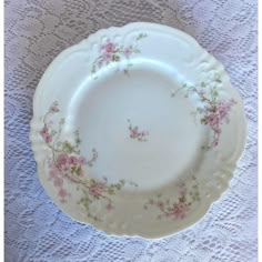 a white plate with pink flowers on it sitting on a lace tablecloth covered table