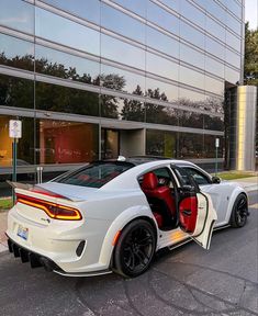 a white sports car parked in front of a glass building with its door open and the driver's seat up