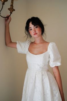 a woman in a white dress is holding a light fixture above her head and looking at the camera