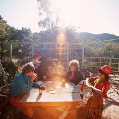 group of people sitting around a table outside on a sunny day with sun shining through the trees