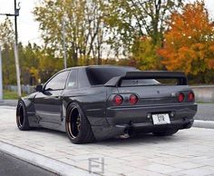 the rear end of a black sports car parked in front of a parking lot with trees