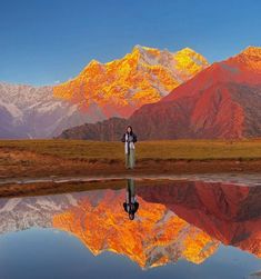 two people standing on the edge of a lake in front of mountains with their backs turned to the camera