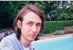 a man with long hair standing in front of a swimming pool and looking at the camera