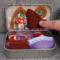 someone is holding their finger in a tin with miniature houses and mushrooms on it's side