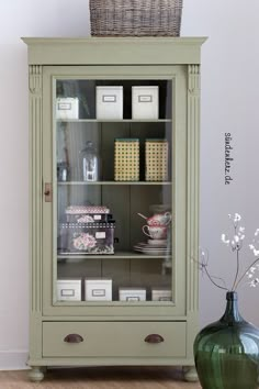 a green china cabinet with drawers and baskets on top, next to a vase filled with flowers