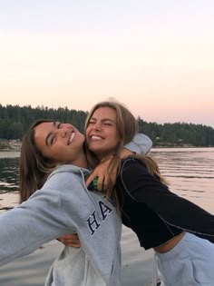 two young women standing next to each other in front of a body of water with trees in the background