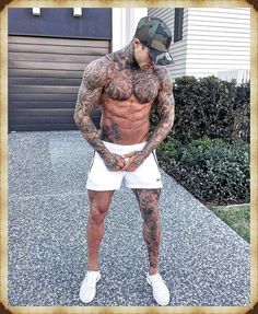 a man with tattoos and no shirt standing in front of a house wearing white shorts