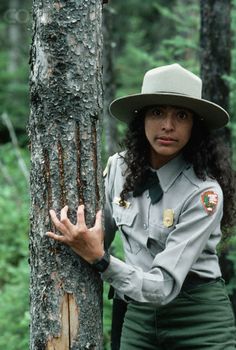 a woman wearing a hat standing next to a tree in the woods with her hands on it