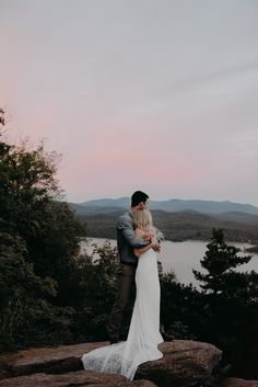 a bride and groom embracing on top of a mountain overlooking the lake at sunset with mountains in the background