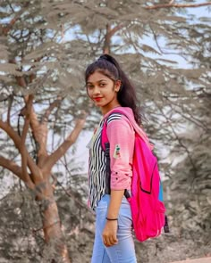 a girl with a pink backpack and blue jeans is standing in front of some trees