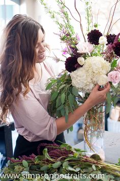 a woman arranging flowers in a vase at a table with greenery and other items