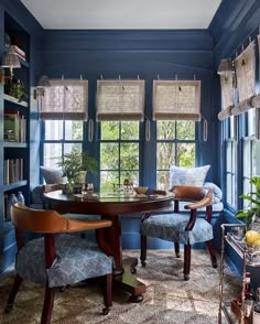 a dining room with blue walls and wooden chairs around a table in front of two windows