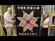 two people holding up a colorful quilt in front of a bookcase full of books