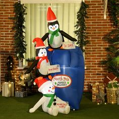 an inflatable penguin and two snowmen with holiday signs on their heads, sitting next to a christmas tree