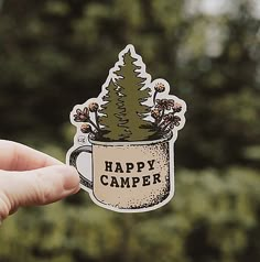 a hand holding up a sticker with the words happy camper on it in front of trees