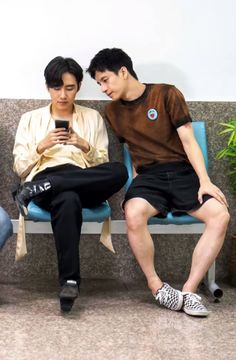 two young men sitting on a bench looking at their cell phones while one looks at his phone