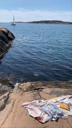 there is a blanket on the rocks by the water with a sailboat in the distance