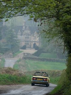 A car going down a road that leads to a beautiful cream colored brick house. The house is old money style with green gardens and bridges that goes over a  small river. Large House, Country Road, Sports Car, Road, Sports, Yellow