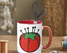 a red and white coffee mug with scissors on it next to thread spools