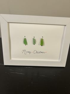 two green christmas trees in a white frame on a black table with the words merry christmas written below them