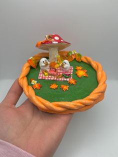 a hand is holding a small toy mushroom on a green tray with leaves and mushrooms