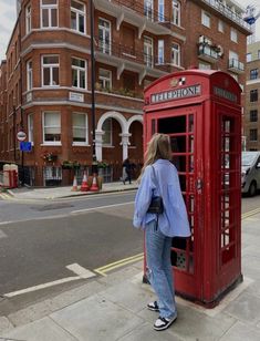 a woman standing next to a red phone booth on the side of a road in front of a tall brick building