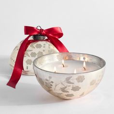 two lit candles in a glass bowl with a red ribbon around the edge and an ornament behind it