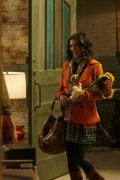 a woman in an orange coat and plaid skirt carrying flowers while walking into a building