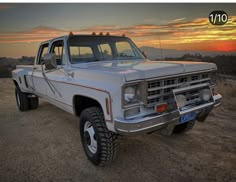 a white pickup truck parked on top of a dirt field at sunset with the sun setting in the background