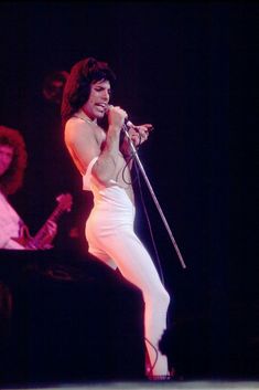 a man in white pants holding a microphone on stage with two other people behind him