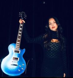 a woman holding up a blue guitar in front of a dark background with her hand on the neck