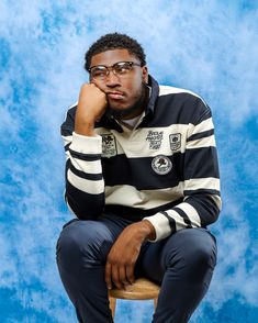 a man sitting on top of a stool in front of a blue background wearing glasses