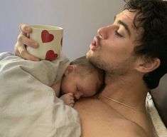 a man laying in bed holding a baby and drinking out of a coffee cup with hearts on it