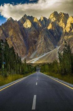 an empty road in the middle of mountains with trees on both sides and clouds above