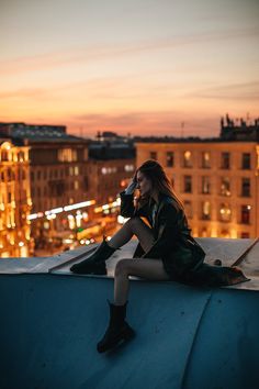 a woman sitting on top of a building next to a tall building at night with the city lights in the background