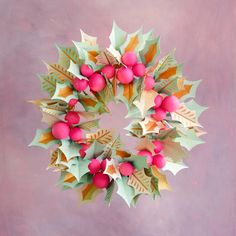 a christmas wreath made out of paper with red balls and green leaves on pink background