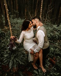 a man and woman are sitting on a swing in the woods, holding onto each other