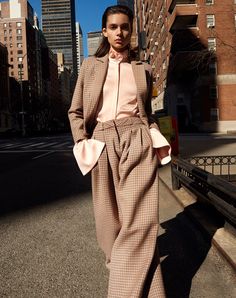 a woman standing on the side of a road in front of tall buildings wearing a pink top and plaid pants