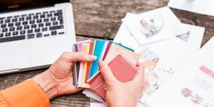 a person is holding several cards in front of a laptop on a wooden table with other papers